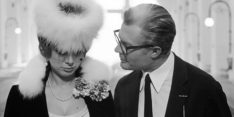 Eine Person mit einer markanten Brille schaut eine andere Person mit einem Hut und einem Schleier an.